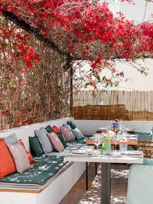 Shady restaurant terrace with a flower rooftop, cosy stone bench with many colourful cushions and set tables.